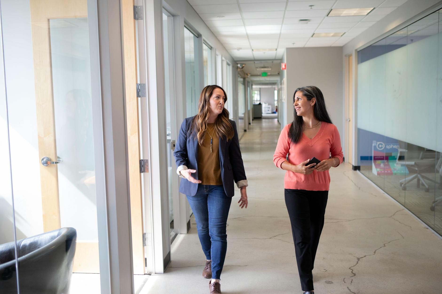Two professional women discussing while walking through a modern office corridor