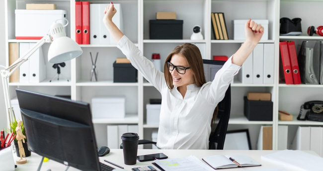 Woman with arms raised in triumph at her home office desk, representing remote work success and digital flexibility