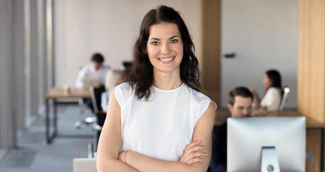 Woman with arms crossed standing in front of colleagues working at computers in a modern office setting