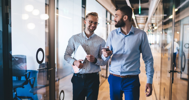 Two male colleagues chatting while walking through a modern office corridor
