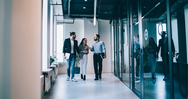 Three coworkers walking and discussing in a bright, glass-walled office hallway