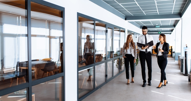 Three formally dressed individuals walking through a corridor, discussing content in an open folder, illustrating collaboration and planning