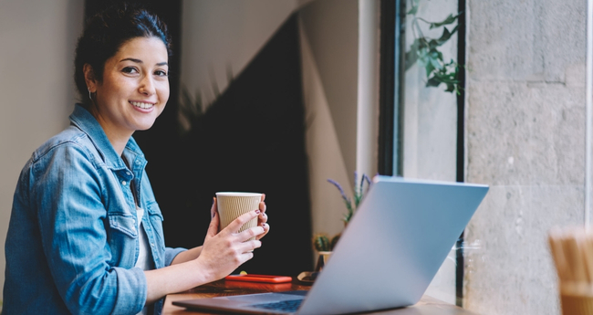 Person holding a coffee cup and smiling while working on a laptop in a relaxed office environment