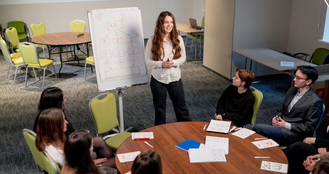Woman presenting to group seated around table with diagrams on whiteboard